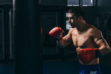 Muscular shirtless athlete in red boxing gloves throwing punch in dark boxing gym. Confident boxer with tattoo on arm doing boxing cardio hitting punch bag. Boxing practice.