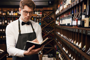 Professional male Sommelier dressed in bowtie and apron writes at notepad, putting down information...