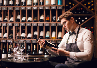 Handsome and young smiling brown haired sommelier with a bottle of wine on the background of dark wine house with shelves of bottles with alcohol