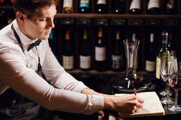 Wine tasting experience in hotel or restaurant. Professional guy or sommelier holding glass of delicious red wine appreciting his tint with bottles collection on background