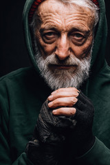 Indoor face portrait of old-aged homeless hobo man dressed in green hoodie with hood on head breathing on hands in diverse mittens to keep them warm at cold day.