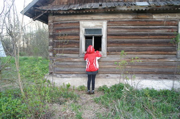 A little girl in a red jacket looks into the window of an old village house.