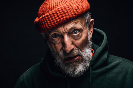 Elderly man indoor studio portrait wearing orange hat, having white beard and wrinkled weathered face, looking at camera with crazy confused expression. Mental Aging Disorders concept