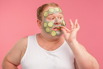 Isolated portrait of funny plump happy man in white undershirt applying cucumber slices over green...