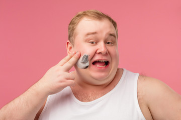 Studio portrait of funny tubby middle-aged man in undershirt smiling at camera with happy joyful expression while shaving his face with razor and foam