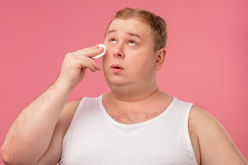 Positive plump casual man doing morning routine cleaning with cotton pads face skin before shaving, isolated over pink background. Grooming concept