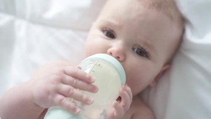 Pretty baby girl drinks milk from bottle lying on bed. Child weared diaper in nursery room.