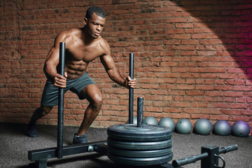 African athletic sportsman pushing sled in cross training gym. Muscular and strong man pushing the...