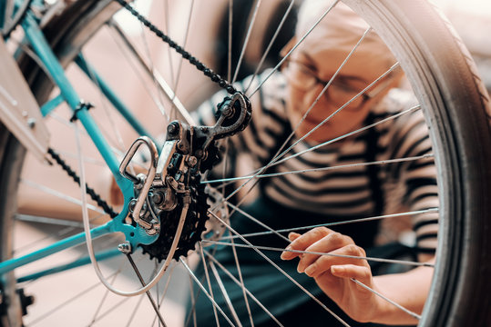 Beautiful Caucasian female worker with short blonde hair and eyeglasses crouching and repairing bicycle. Bike workshop interior.