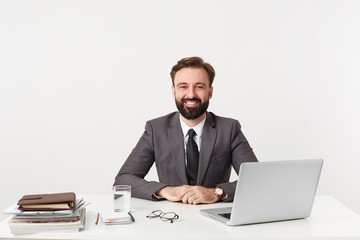 Successful bearded man, businessman, top manager smiling sitting with hands folded on the desktop in office,good-naturedly looks at the camera, dressed in a expensive suit with a tie, has a good watch