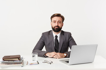 Bearded manager sits with hands folded on the desktop in office, looks at the camera with tired indifferent look, dreams about vacation, dressed in a expensive suit with a tie, has a good watch