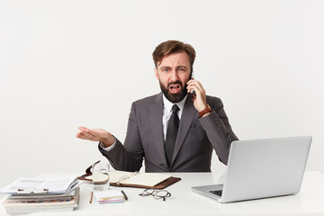 Respectable worthy company manager sitting at desktop, has unpleasant telephone conversation, feels angry, he is told absurd ridiculous nonsensical things, dressed in a suit with a tie, has beard