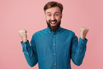 Handsome bearded man suddenly turns out to be a winner, gets a victory, triumphs, happy surprised holds his hands up his fists clenched, overflowing with joy and happiness,over pink background