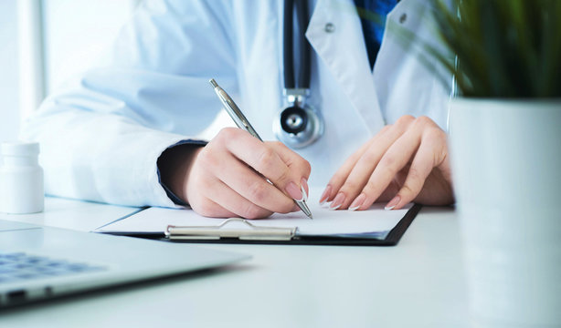 Female doctor filling up prescription form or patient history list at clipboard pad during physical exam or disease prevention while sitting at the desk in hospital closeup.