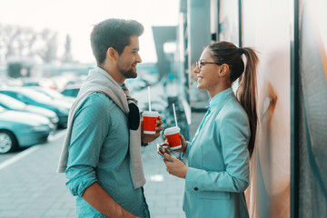 Smiling multicultural couple dressed elegant standing outdoors, looking at each other and drinking coffee to go. Woman holding tablet.