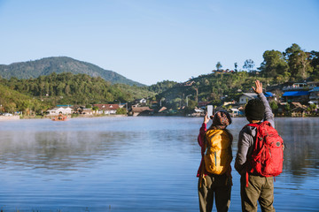 An Asian couple who is standing and watching the fog rising on the lake in the morning, Travel nature. Take a picture of the lake.