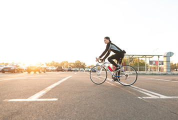 Fototapeta premium Rider in a cap and dark sportswear rides around the city on a white highway, on the background of the sunset. Portrait of a cyclist in motion against the background of the sunset and urban landscape.