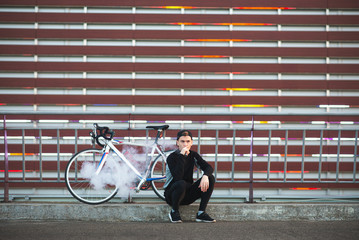 Cyclist sitting bicycle on a striped wall background and smoking a mechanical vape device. Vaper...