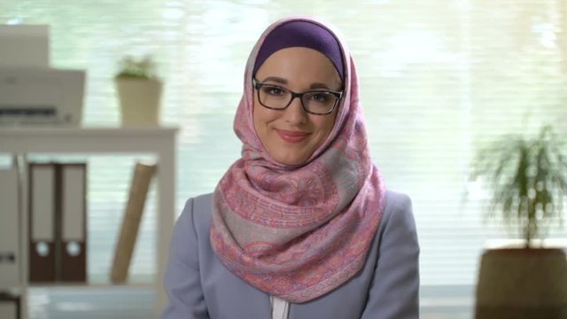 Portrait of professional young muslim business woman looking at camera laughing wearing traditional headscarf in office background close up slow motion