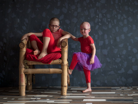 Two funny kids in pink t-shirts and pantyhose playing together in room. One of boys wore lilac skirt. Children with glasses posing for camera next to chair.