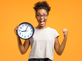 Joyful girl holds clock, raises fist and shouts loudly. Photo of african american girl wears casual...