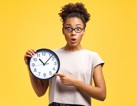 Time to work! Student with shocked face holds clock and points to them. Photo of african american girl wears casual outfit on yellow background. Emotions and pleasant feelings concept.