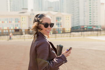 Funny attractive european woman wearing  glasses holding smartphone listening music and walking in the city