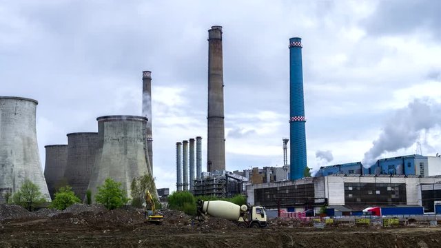 Time lapse of thermal power station with cooling towers emitting smoke, while a few concrete mixer trucks work in the front. Storm clouds moving in. Industrial, energy generation, coal burning.