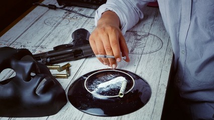 Drugs addicted dealer man's hand holding syringe with cocaine, rolled banknote ,gun, cartridges and black mask on dirty white wooden table floor in his dark room,  crime scene concept
