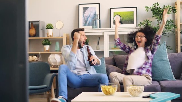 Couple Man And Woman Are Watching Sports On TV Supporting Team Then Doing High-five And Hugging Enjoying Victory. Happy People Are Wearing Casual Clothing And Holding Drinks.