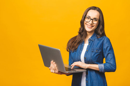 Young Happy Smiling Woman In Casual Clothes Holding Laptop And Sending Email To Her Best Friend. Isolated Against Yellow Background.