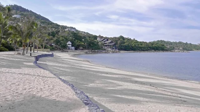 locked wide shot of sandy beach with a jungle mountain shore line in the background, resort and villa buildings in between the paln trees, sunny blue sky day with random clouds.