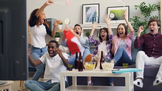 Young Canadians men and women are watching hockey game on TV cheering with flags doing high-five and hugging sitting on sofa at home. Sports and youth concept.