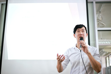 Portrait of young handsome Asian male speaker publicly speaking on stage to group of audience with...