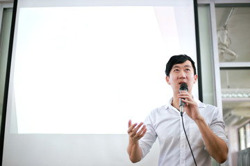 Portrait of young handsome Asian male speaker publicly speaking on stage to group of audience with white board behind - Powered by Adobe