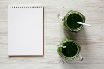 Glass jars full of green smoothie, blank notebook over white wooden surface, top view. Flat lay, overhead, from above. Copy space. - Powered by Adobe