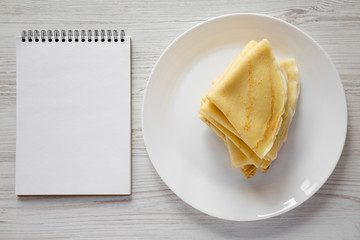 Pile of tasty blini on a white plate, blank notepad over white wooden background, top view. Flat...