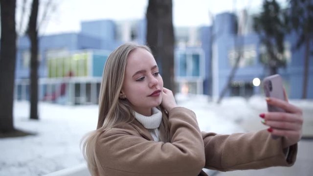 Close up of the pretty young blonde girl taking selfie photos on the smartphone device camera and posing to it. Outdoor.