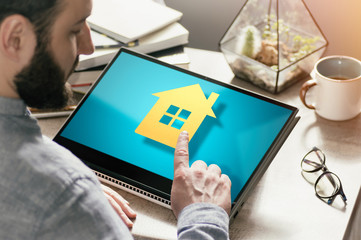 Bearded man with laptop at his desk. He presses on the house icon. Concept of real estate buying, booking, advertising via internet. Image