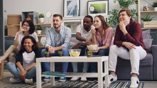 Girls and guys attractive young people are enjoying comedy on TV laughing and eating popcorn sitting on sofa at home. Entertainment and lifestyle concept.