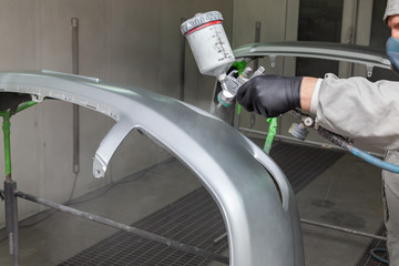 A male worker paints with a spray gun a part of the car body in silver after being damaged at an accident. Plastic elements from the vehicle during the repair in the workshop auto service.