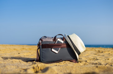 Shoulder bag with a smartphone and a white hat on the sand of the beach