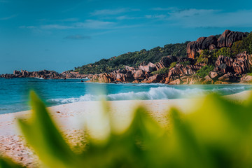 Ocean waves on Grand Anse tropical beach in La Digue, Seychelles. Photo framed by green foliage focusing to famous granite rock formations in background