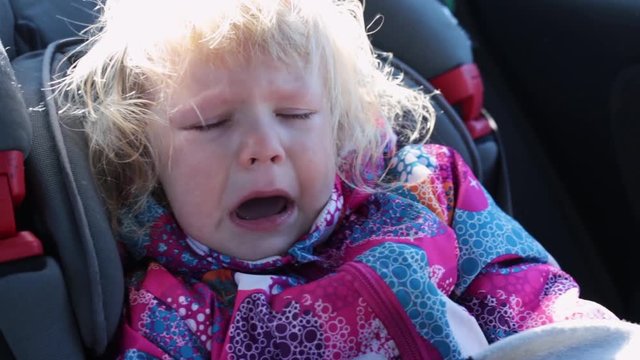 Small Caucasian Child Girl With Curly Hair In A Warm Jacket Sits In A Child Seat, Wearing A Seatbelt Riding In A Car Crying Loudly Tears Running Down Her Cheeks.