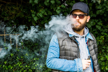 man with a beard smokes an electronic cigarette in a park