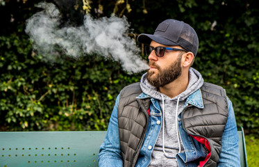 man with a beard smokes an electronic cigarette sit on a bench