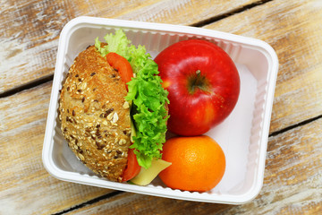 Healthy lunchbox containing brown cheese roll with lettuce and tomato, apple and orange