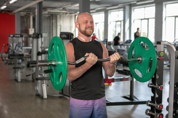 Obraz na płótnie Canvas Bald athlete in a black T-shirt and blue shorts with headphones, lifting the barbell with effort. He is standing in the gym and smiling