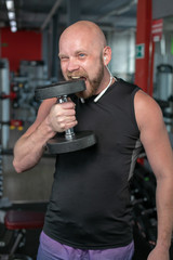 Fototapeta na wymiar bald man bites his teeth into an iron dumbbell in the gym. Sportsman with a dumbbell in mouth
