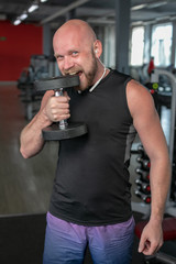 Fototapeta na wymiar bald man bites his teeth into an iron dumbbell in the gym. Sportsman with a dumbbell in mouth